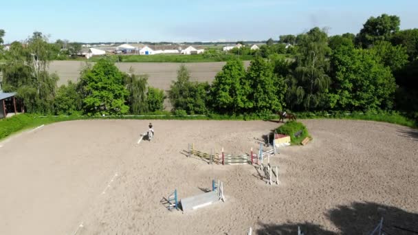 Vista desde arriba, filmación de video aéreo, campo de arena de entrenamiento, parque infantil, jinetes, jinetes montan caballos, realizan varios ejercicios con caballos, junto a las barreras. verano, al aire libre , — Vídeos de Stock