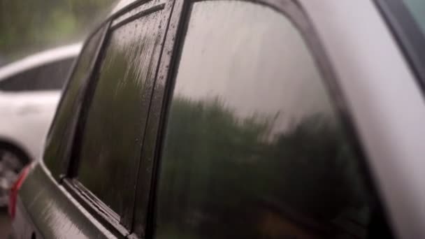 Vista desde el exterior, hay lluvia fuerte, ducha, gotas caen sobre ventanas teñidas de coche negro vacío de pie en el estacionamiento. gotitas fluyen por el vidrio en pequeños arroyos — Vídeo de stock