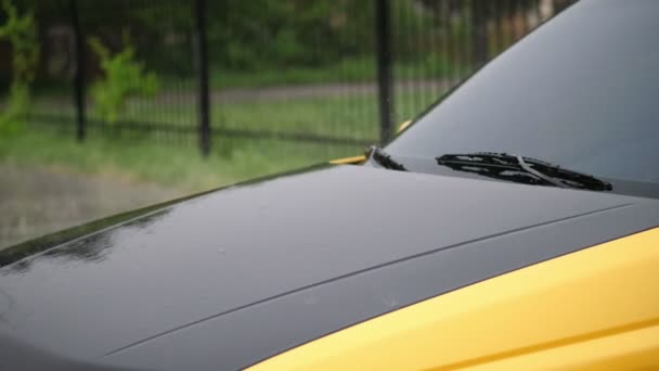 Vista desde el exterior, hay lluvia fuerte, ducha, gotas caen con salpicaduras de agua, en la capucha del coche mojado, parabrisas con limpiaparabrisas de coche vacío negro y amarillo de pie en el estacionamiento — Vídeos de Stock