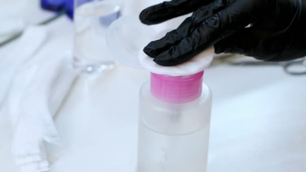 Nahaufnahme, die Hände der Kosmetikerin in schwarzen Gummihandschuhen halten das Wattepad und befeuchten es mit einem Spender in einer speziellen transparenten Flüssigkeit aus der Flasche. — Stockvideo