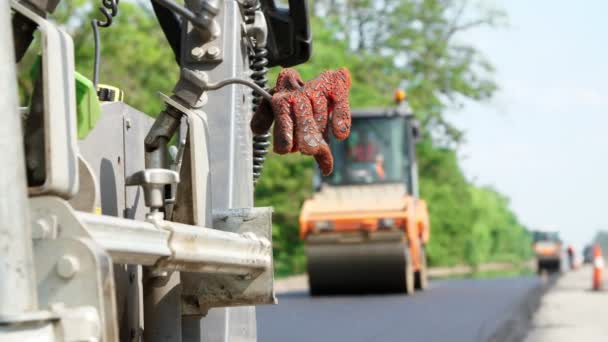 公路、公路建设工程的维修。在前景上, 在铺设沥青的机器上, 工人留下了特殊的橙色手套。沥青混凝土修整器铺设新的沥青路面 — 图库视频影像