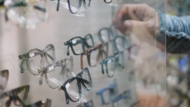 Närbild, showcase i optiska store, optik, optiker butik, Glasögon butik, mycket glas, ramar, mans hand väljer fashionabla korrigerande glasögon, Glasögon . — Stockvideo