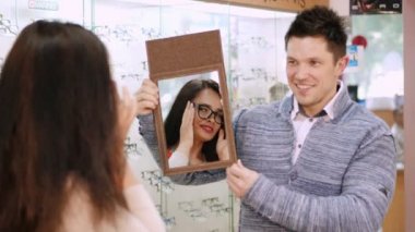 seksi güzel kadın yeni gözlüklerini optik mağaza, optik, gözlükçü perakende mağaza, gözlük dükkanı, bir ayna tutan yakışıklı genç erkek satıcısı bir aynadaki kontrol