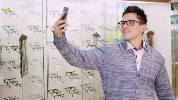 Σέξι όμορφος νεαρός άνδρας με γυαλιά, γυαλιά κάνει selfie σε οπτικό κατάστημα οπτικών, οπτικό κατάστημα λιανικής πώλησης, κατάστημα γυαλιών, ο άνθρωπος κοιτάζει την κάμερα μέσα mobilephone του — Αρχείο Βίντεο