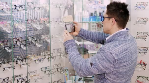 Bonito homem em óculos tirando fotos para óculos na loja de óptica, óptica, loja de varejo de óptica, loja de óculos, para ver mais tarde em casa que modelo comprar, ou compartilhar redes sociais — Vídeo de Stock