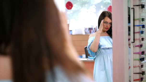 Молодая сексуальная женщина проверяет свои новые очки в зеркале в магазине оптики. Молодая женщина в очках в магазине оптики. Красивая девушка в очках в магазине оптики — стоковое видео