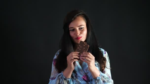 Retrato de una mujer morena sexy con labios rojos que erótica y juguetonamente come chocolate negro con avellanas y mirando a la cámara en el estudio. fondo gris oscuro . — Vídeos de Stock