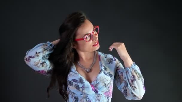 Retrato de una mujer morena sexy con labios rojos que erótica y juguetonamente se prueba gafas con estilo, gafas y mirando sexualmente a la cámara, posando en el estudio. fondo gris oscuro . — Vídeos de Stock