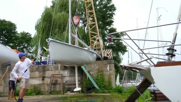 Κίεβο, Ουκρανία, Yachting-Ιούλιος 29, 2018: στην όχθη ποταμού Δνείπερου, στην προβλήτα, ανύψωσης γερανού εγείρει λευκό γιοτ, ο άνθρωπος στο ειδικό ρούχα, με ειδική σφουγγαρίστρα, μηχανή πλένει το γιοτ — Αρχείο Βίντεο