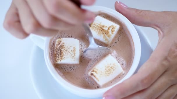 Gros plan, vue sur le dessus, tasse blanche avec cappuccino, avec mousse crémeuse épaisse et guimauves. une main de femme agite lentement la boisson avec une cuillère à café — Video