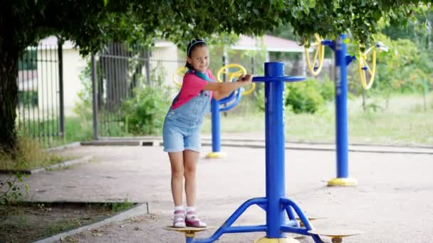 Sonriente, feliz niña de ocho años comprometida, haciendo ejercicios en equipo de ejercicio al aire libre, al aire libre, en el parque, verano, día caluroso durante las vacaciones . — Vídeos de Stock