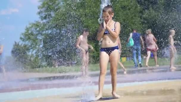 Sorridente, felice bambina di otto anni in costume da bagno divertirsi in spruzzi in fontana di città di strada, all'aperto, nel parco, estate, calda giornata di sole durante le vacanze . — Video Stock