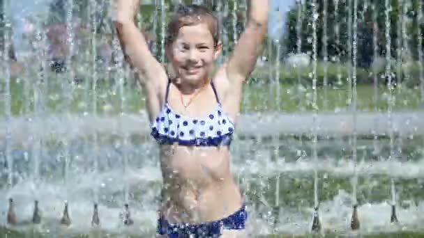 Улыбаясь, счастливая восьмилетняя девочка в купальнике веселится в брызгах в фонтане уличного города, на открытом воздухе, в парке, летом, жаркий солнечный день во время отпуска . — стоковое видео