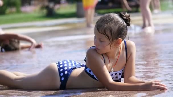 Smiling, happy eight year old girl in swimsuit having fun in splashes in street city fountain, outdoors, in park, summer, hot sunny day during vacation. — Stock Video