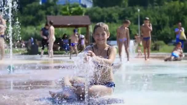 Усміхнена, щаслива восьмирічна дівчина в купальнику розважається в бризках на вулиці міський фонтан, на відкритому повітрі, в парку, влітку, гарячий сонячний день під час відпустки . — стокове відео
