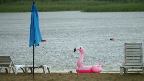Летний дождь, гроза, ливень на пляже, у реки. одинокие пустые шезлонги и пляжные зонтики стоят, кто-то забыл надувной круг в виде розового фламинго . — стоковое видео