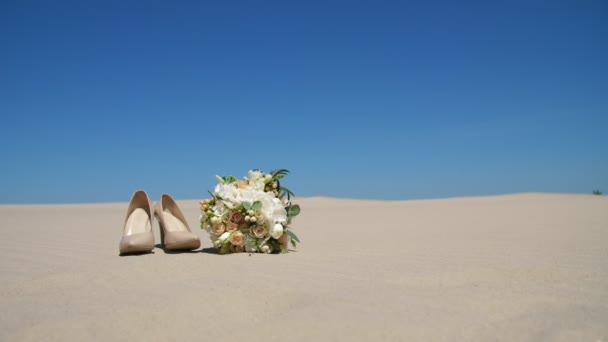 Літо, пустеля, весільні жіночі бежеві підбори стоять на піску, поруч з весільним букетом. День, на тлі блакитного неба . — стокове відео