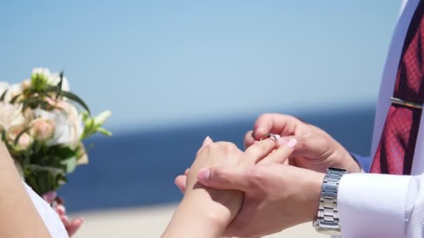 Close-up, a noiva eo noivo usar anéis uns aos outros, contra o fundo do mar, areia e céu azul. verão, dia . — Vídeo de Stock