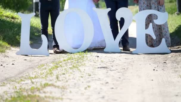 Auf einem Feldweg stehen große weiße Buchstaben für Liebe, vor dem Hintergrund der Füße eines Hochzeitspaares und Brautjungfern und Freund des Bräutigams. Sommer, Tag, Hochzeitsdekor. — Stockvideo