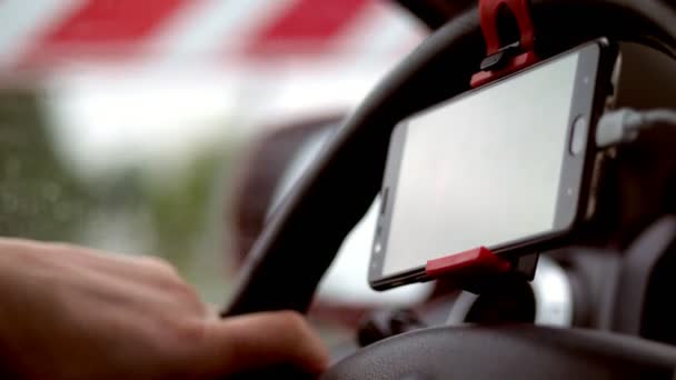 Primer plano, en el coche, la mano del conductor sostiene el volante del coche, un gadget, teléfono móvil, navegador está conectado al volante . — Vídeos de Stock