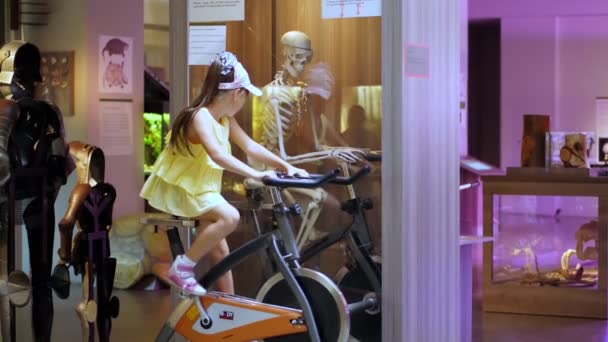 Cheerful attraction. The girl twists pedals of bicycle, holds the steering wheel with both hands. and next to, behind the glass on bike sits a skeleton and also turns the pedals of a bicycle. — Stock Video