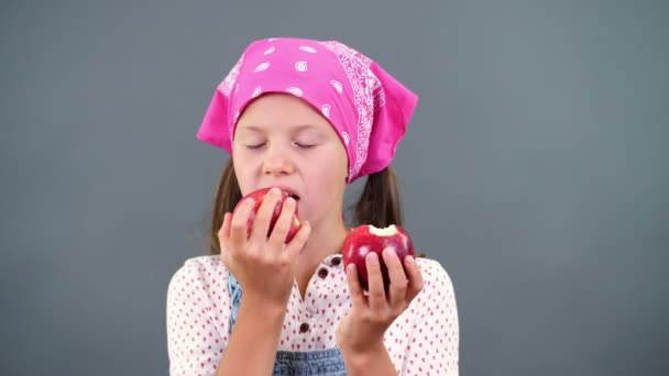 Aranyos mezőgazdasági termelő girl gazdaság a kezében két nagy érett piros alma, eszik, harap őket egy szürke háttér, a stúdióban. Egészséges táplálkozás koncepció. — Stock videók