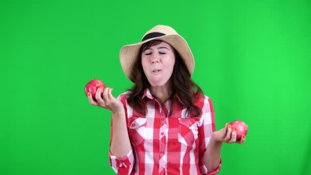 Engraçado, sorridente agricultor feminino em camisa xadrez e chapéu, segurando nas mãos duas grandes maçãs azedas, come, morde-los, rugas seu rosto, no fundo verde no estúdio. Comida saudável à sua mesa . — Vídeo de Stock