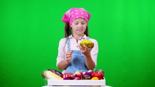 Bonito, sorrindo, pequena menina agricultor mostra uma caixa com diferentes vegetais frescos, colheita. sobre fundo verde no estúdio. Comida saudável à sua mesa, nutrição saudável . — Vídeo de Stock