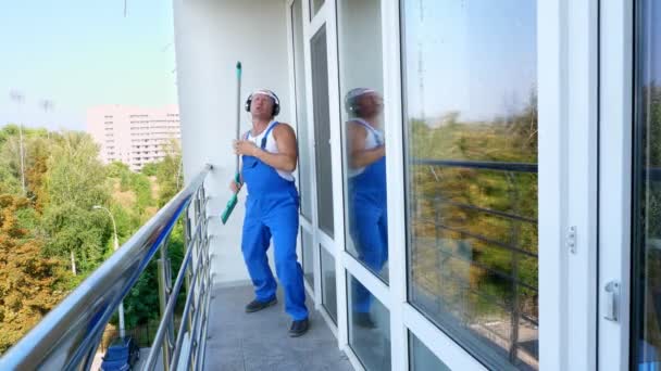 Kaukasischer attraktiver Mann, Reinigungskraft, in blauen Overalls mit Kopfhörern, hat Spaß, tanzt, spielt auf Fensterwischgitarre, fröhlich und verrückt — Stockvideo