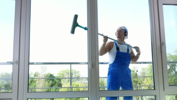 Вид через окно, красивый мужчина работник уборки, в синем комбинезоне, с наушниками,, Чистка, мытье окон специальной шваброй, наслаждаясь своей работой и музыкой . — стоковое видео