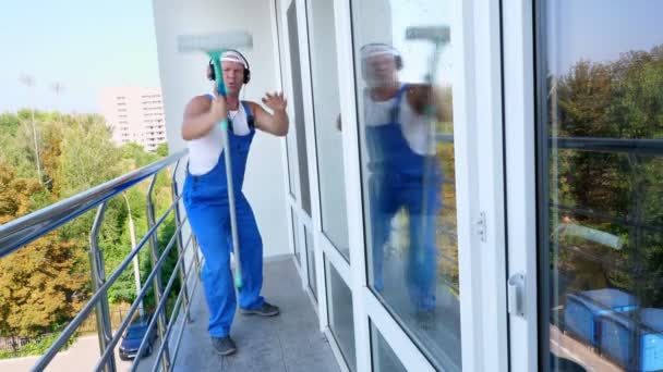 Kaukasischer attraktiver Mann, Reinigungskraft, in blauen Overalls mit Kopfhörern, hat Spaß, tanzt, spielt auf Fensterwischgitarre, fröhlich und verrückt — Stockvideo