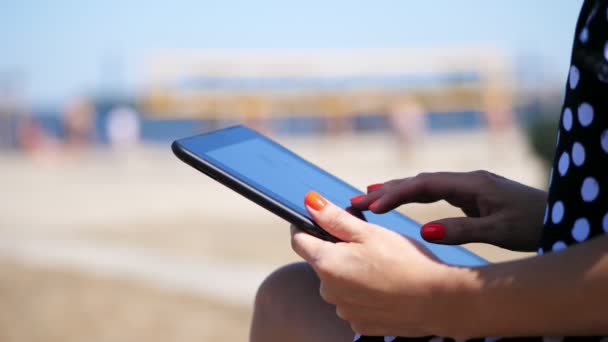 Primer plano, manos femeninas con manicura de color rojo brillante, chica trabaja en una tableta, en la playa, en un día caluroso de verano, sobre un fondo de voleibol de playa — Vídeo de stock