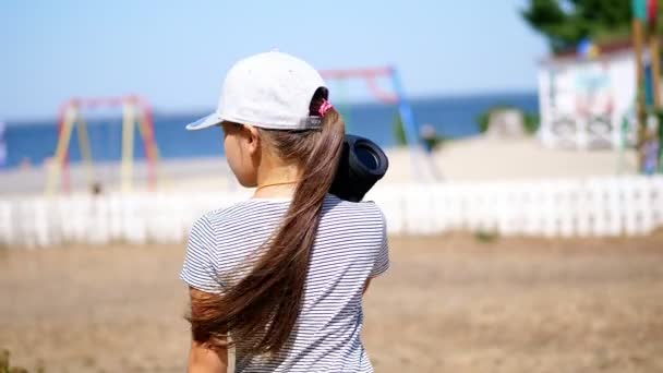 Vista traseira, criança adolescente, menina loira de cabelos longos em um blazer ouvir música com alto-falante portátil bluetooth, na praia, dançando, em um dia quente de verão . — Vídeo de Stock