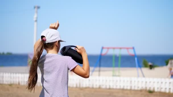 Vista traseira, criança adolescente, menina loira de cabelos longos em um blazer ouvir música com alto-falante portátil bluetooth, na praia, dançando, em um dia quente de verão . — Vídeo de Stock