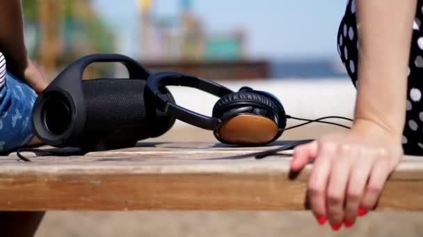 Nahaufnahme, große Kopfhörer und ein kleiner, tragbarer, schwarzer Bluetooth-Mini-Musik-Lautsprecher auf der Bank, im Sommer am Strand — Stockvideo