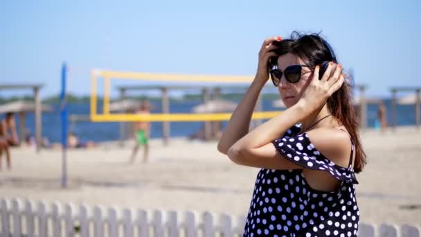 Портрет, красивая девушка в солнечных очках в больших черных наушниках, слушающая музыку со смартфона, на пляже, в жаркий летний день, на фоне пляжного волейбола — стоковое видео