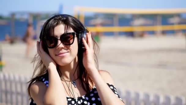 Портрет, красива дівчина в сонцезахисних окулярах в великих чорних навушниках, слухаючи музику зі смартфона, на пляжі, в спекотний літній день, на фоні пляжного волейболу — стокове відео