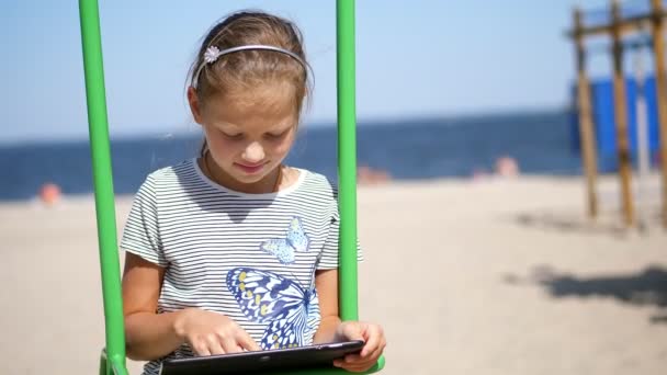 Porträt, Teenager-Kind, langhaariges blondes Mädchen, arbeitet an einem Tablet, am Strand, an einem heißen Sommertag, vor dem Hintergrund des Meeres — Stockvideo