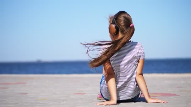 バック ビュー、十代の子、海の近くの暑い夏の日にビーチでのスマート フォンからの音楽を聴いて、大きなヘッドホンを長い髪の金髪少女 — ストック動画