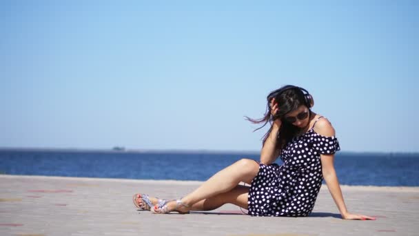 Linda menina morena em óculos de sol e vestido de sol, com grandes fones de ouvido, ouvindo música do smartphone, na praia, no dia quente de verão, perto do mar — Vídeo de Stock