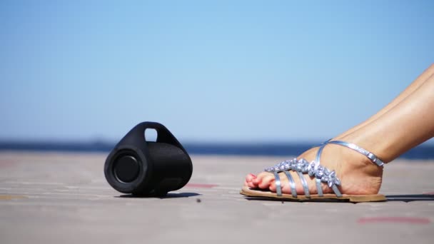 Крупный план, мини-музыка Bluetooth портативный черный цилиндр беспроводной громкоговоритель. женские ноги в серебряных сандалиях, с ярко-красным педикюром, танцующие под музыку. на пляже, летом — стоковое видео