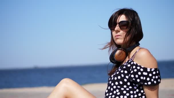 Portrait, belle brune, femme en lunettes de soleil, et écouteurs, assis sur la plage, contre la mer. vent fait onduler ses cheveux — Video