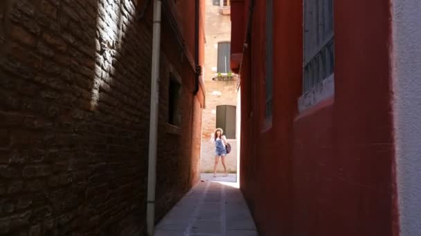 VENECIA, ITALIA - 7 DE JULIO DE 2018: al final de una calle muy estrecha de Venecia, una joven turista, con sombrero y mochila, se para y mira, examina la arquitectura . — Vídeos de Stock