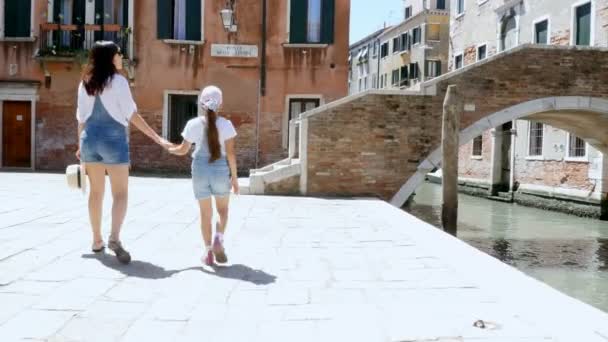 VENECIA, ITALIA - 7 DE JULIO DE 2018: una mujer joven y una niña en monos de mezclilla idénticos, pantalones cortos, caminar por las calles de Venecia, subir a un pequeño puente sobre el canal. verano día caliente . — Vídeos de Stock