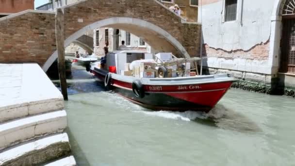 VENICE, ITALIEN - JULI 7, 2018: langs en smal kanal, under en bro, passerer fragtbåde, leverer alt, hvad der er nødvendigt i byen. sommer varm dag . – Stock-video