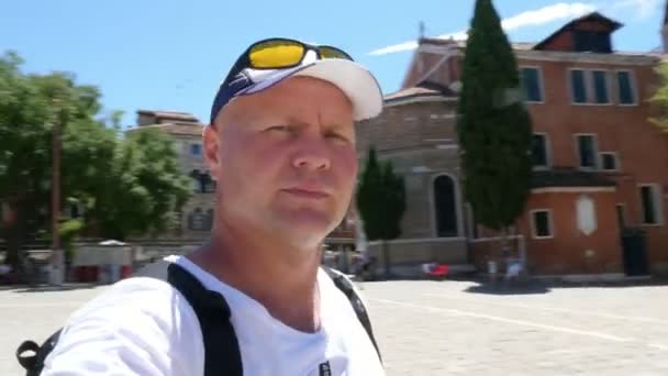 VENICE, ITÁLIA - JULHO 7, 2018: Um turista masculino tira vídeos selfie na Piazza Venezia. Mulher engraçada em óculos de sol e chapéu está se mexendo, se divertindo no fundo. Turistas em Veneza . — Vídeo de Stock