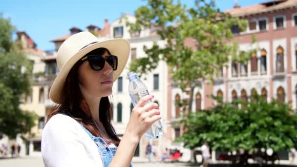 ВЕНИЦА, ИТАЛИЯ - 7 июля 2018 года: Молодая женщина в солнцезащитных очках и шляпе пьет чистую альпийскую воду из бутылки, на фоне архитектуры Венеции, в жаркий летний день . — стоковое видео