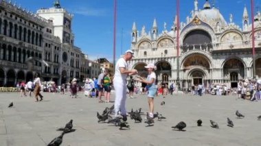 Venice, İtalya - 7 Temmuz 2018: mutlu bir adam ve çocuk kız, turistler, besleme, güvercin, oyun ile onları, holding görünümünü Piazza San Marco, St Marks Bazilikası, bir yaz gününde eğleniyor