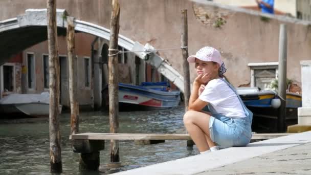 Benátky, Itálie - 7 července 2018: smutná dívka dítě, v kraťasech a čepici se nachází na břehu malého vodního kanálu, poblíž mostu v Benátkách. horký letní den. — Stock video