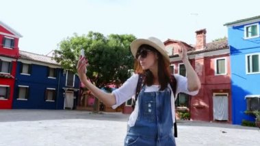 Venedik, Burano, İtalya - 7 Temmuz 2018: turist, Genç kadın, güneş gözlüğü, şapka, sırt çantası yapar smartphone ile selfie karşı Burano Adası'nın çok renkli evleri 